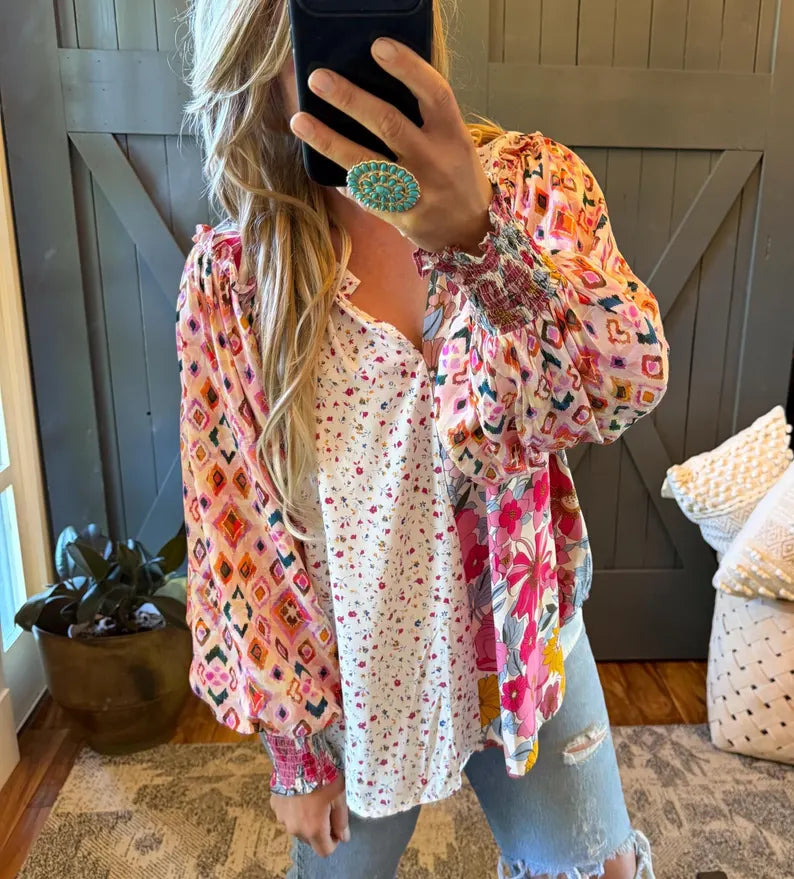 A woman takes a mirror selfie wearing a layered bohemian-style outfit featuring a white long-sleeve top with tiny red and blue dots, a pink and orange patterned cardigan with diamond motifs, and light blue distressed jeans.