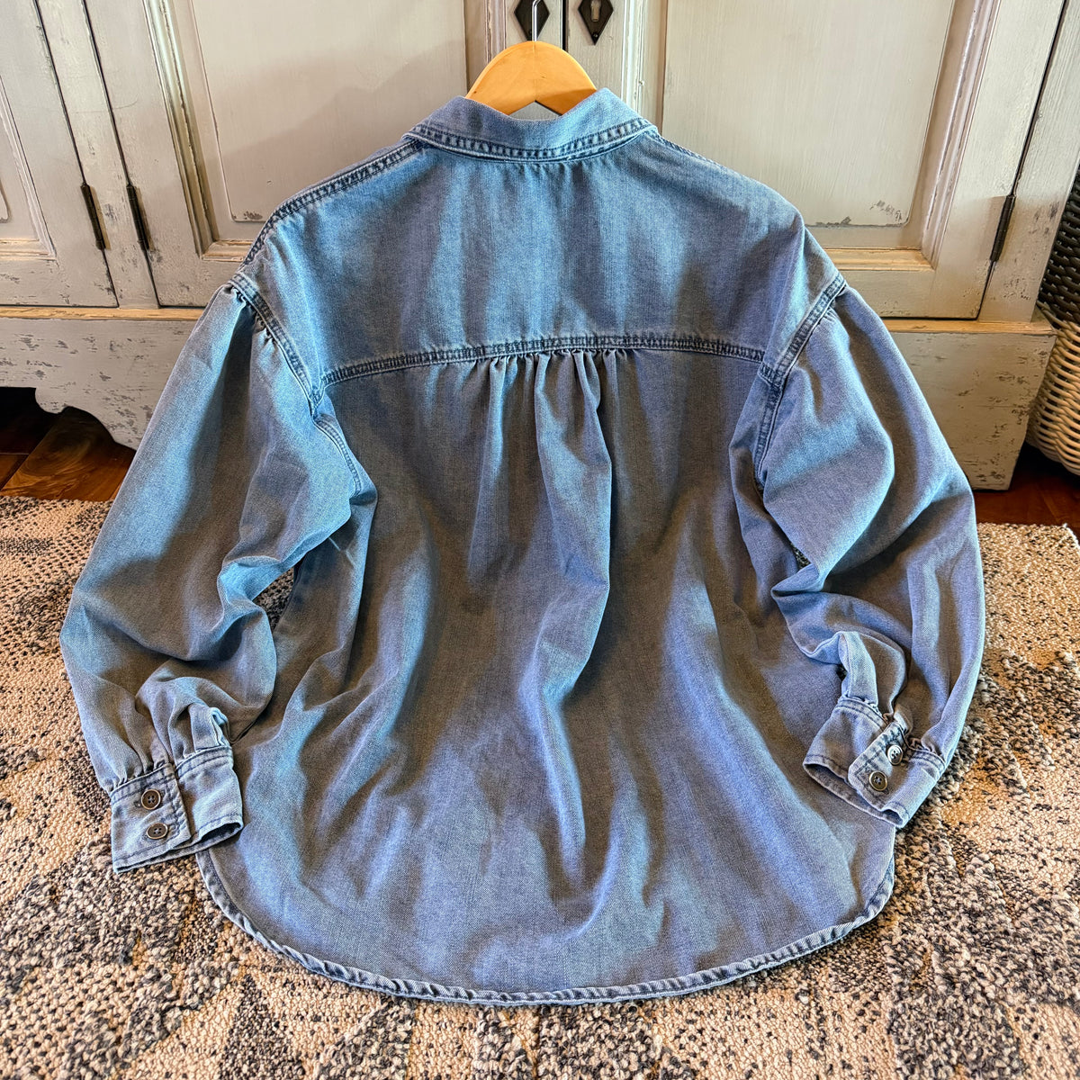 A light blue denim button-up shirt with long sleeves and a gathered back seam, hanging on a wooden hanger on a patterned rug in front of a distressed white cabinet.
