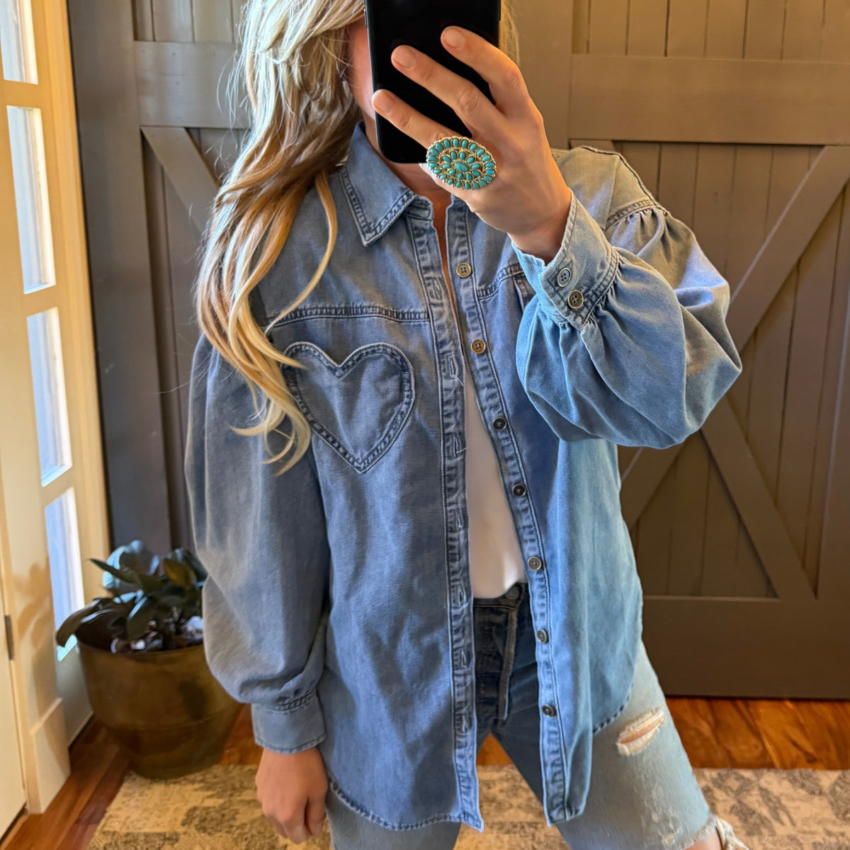 A woman takes a mirror selfie wearing an oversized light-wash blue denim shirt with a heart-shaped chest pocket, paired with distressed jeans and a white top. She is holding a black smartphone in front of her face.