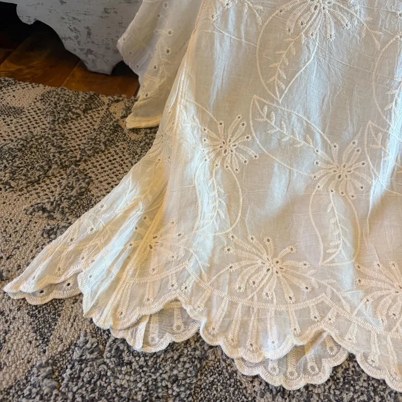 An off-white embroidered cotton fabric with floral and leaf patterns and a scalloped hem, draped over a textured gray rug.