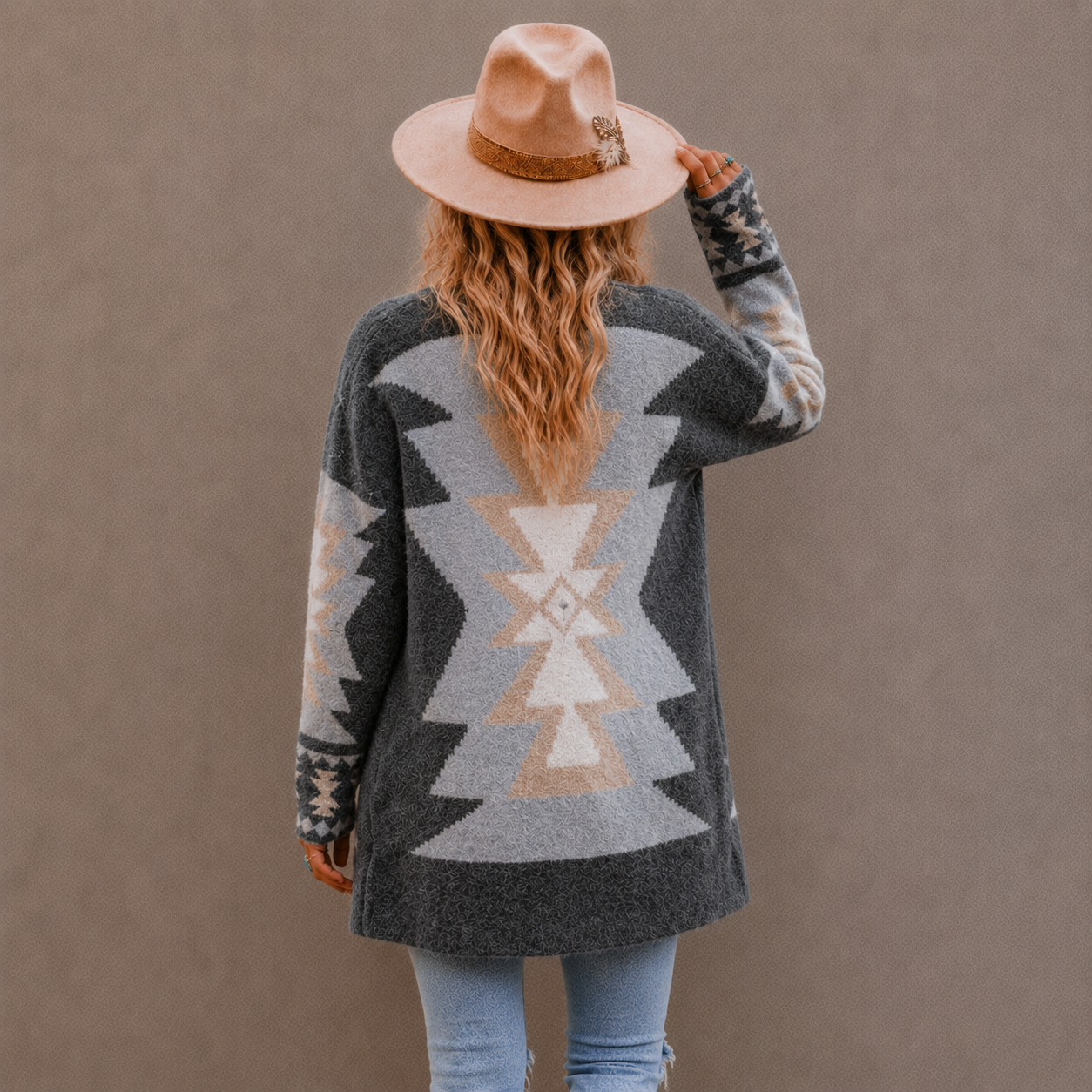 A woman viewed from behind wearing a dark gray knit cardigan with a large beige and light gray geometric Southwestern-style pattern, a tan hat, and light blue jeans, standing against a solid taupe background.