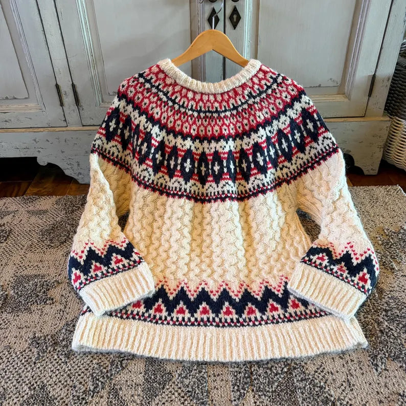 A cream-colored cable-knit sweater with red and navy blue Fair Isle patterns, hanging on a wooden hanger on a patterned rug in front of a rustic white cabinet.