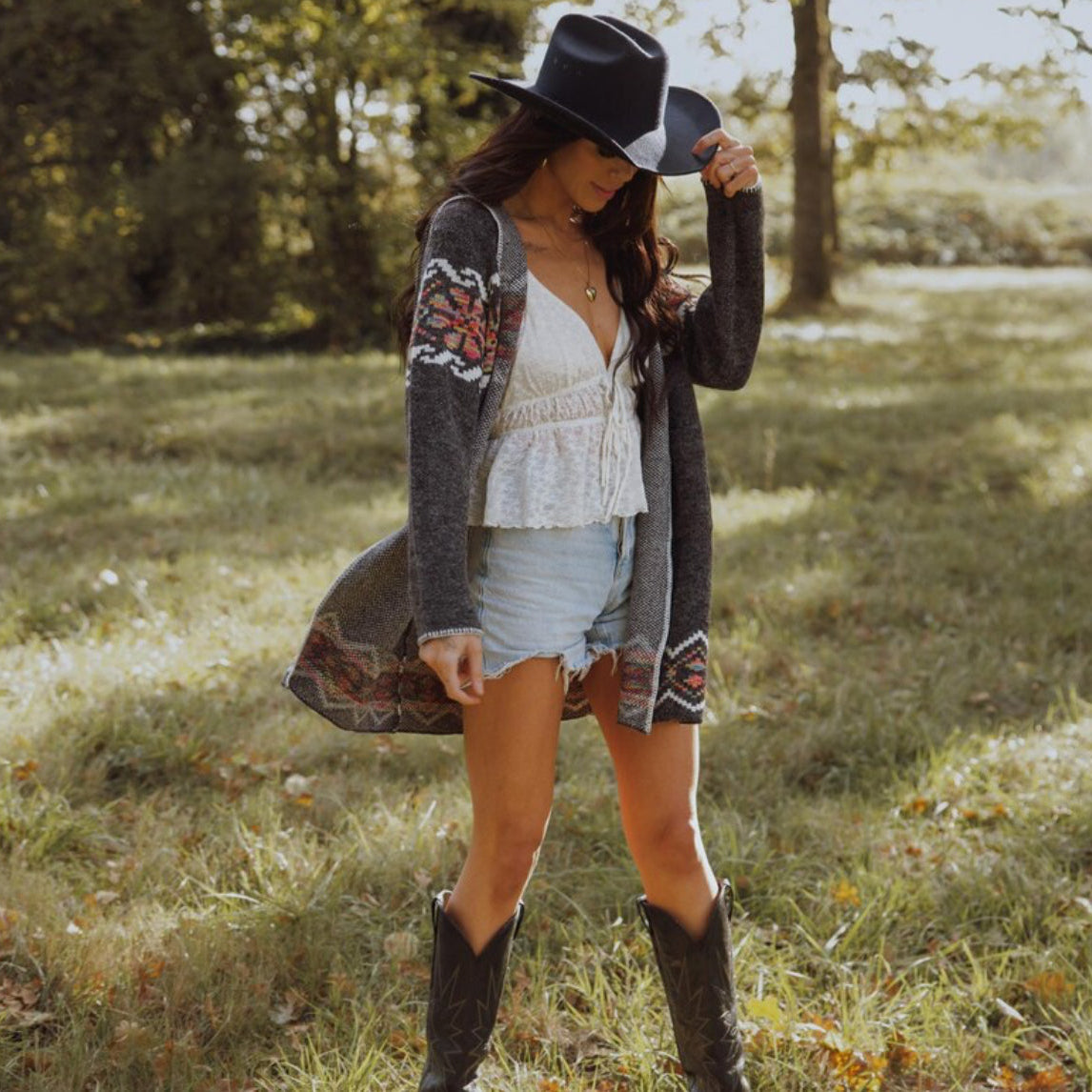Woman in a casual outfit with a cardigan, shorts, and boots standing in a grassy field.