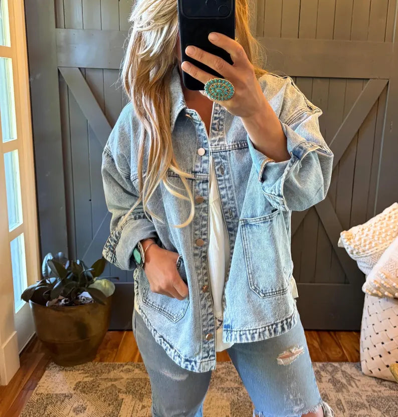 A woman takes a mirror selfie wearing an oversized light-wash blue denim shirt jacket over a white top and distressed blue jeans. She is holding a black smartphone, with a green stone ring on her finger, in front of a gray wooden barn-style door.