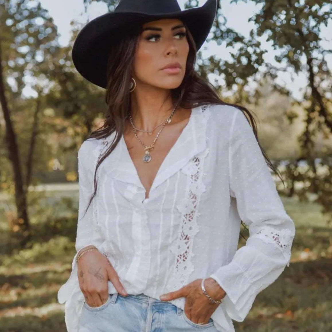 Woman wearing a white blouse with lace details and a black cowboy hat in a natural setting.