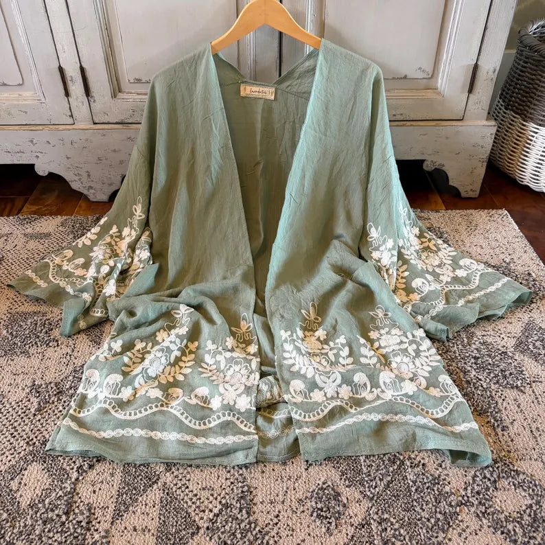 A sage green open-front kimono cardigan with wide bell sleeves and white floral embroidery, hanging on a wooden hanger on a patterned rug.