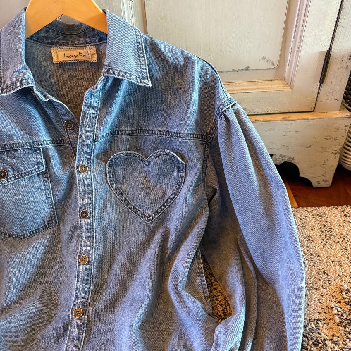 A light blue denim shirt jacket with a heart-shaped chest pocket, hanging on a wooden hanger against a rustic white wooden door and speckled tile floor.