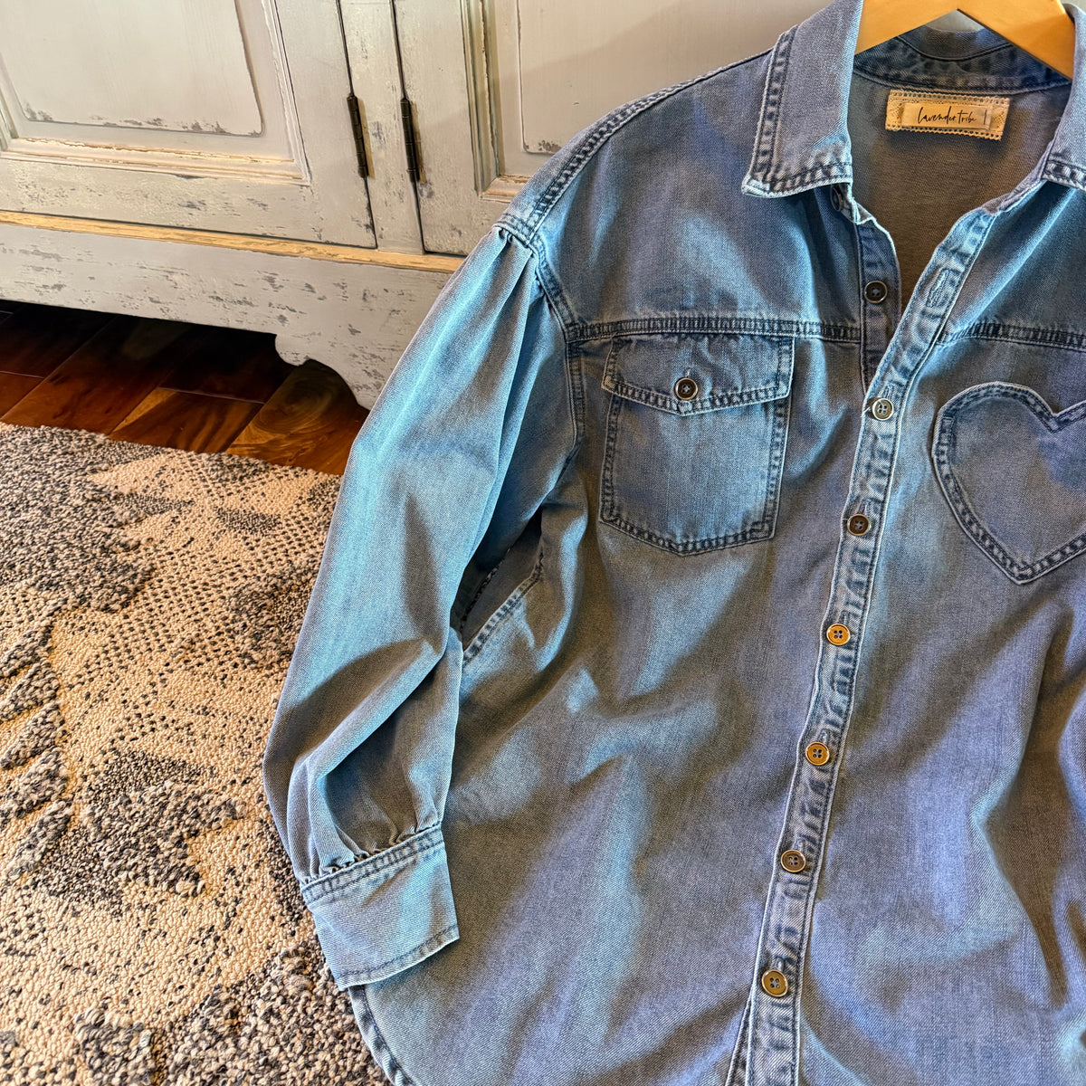 A light blue denim button-up shirt with a heart-shaped chest pocket, laid flat on a patterned rug in front of a distressed white cabinet.