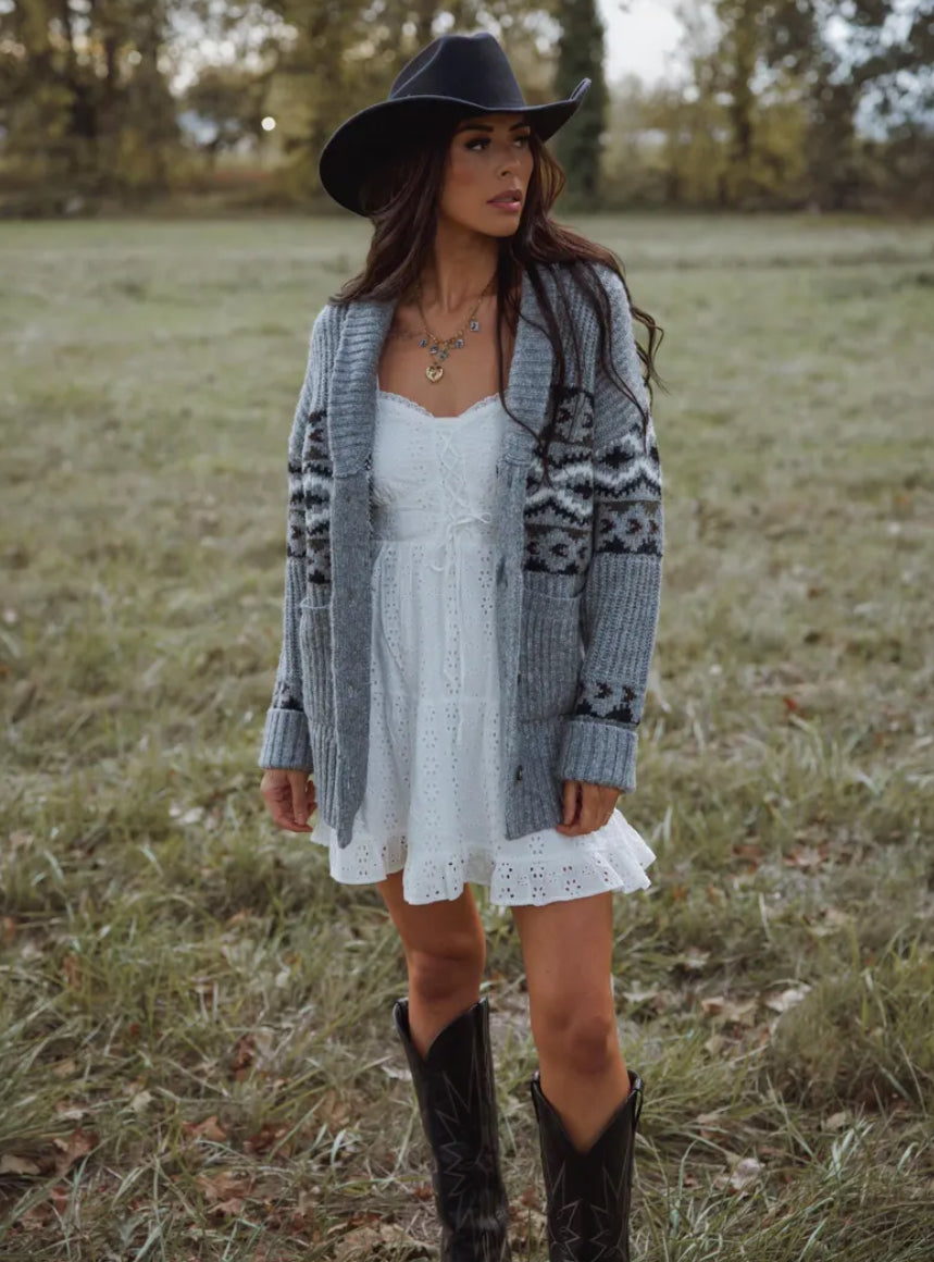 Woman in a white dress, gray cardigan, and black boots standing in a field.