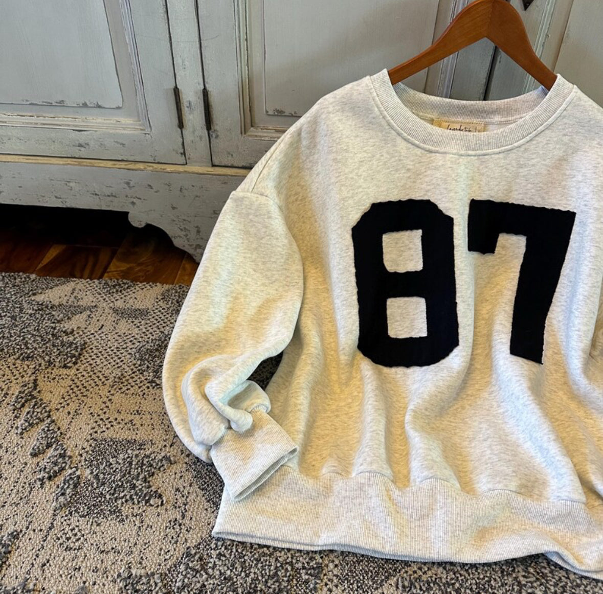 White sweatshirt with black number 87 on a hanger against a wooden cabinet background