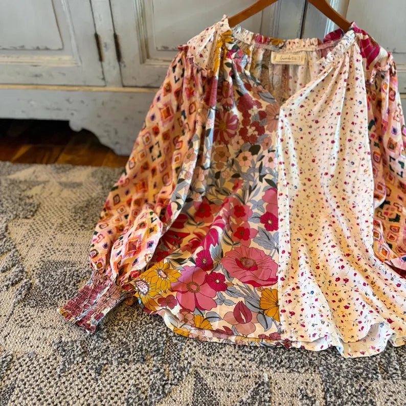 A bohemian-style long-sleeve blouse with mixed prints, including a pink floral pattern and a small red polka dot print on a cream background, hanging on a wooden hanger over a patterned rug.