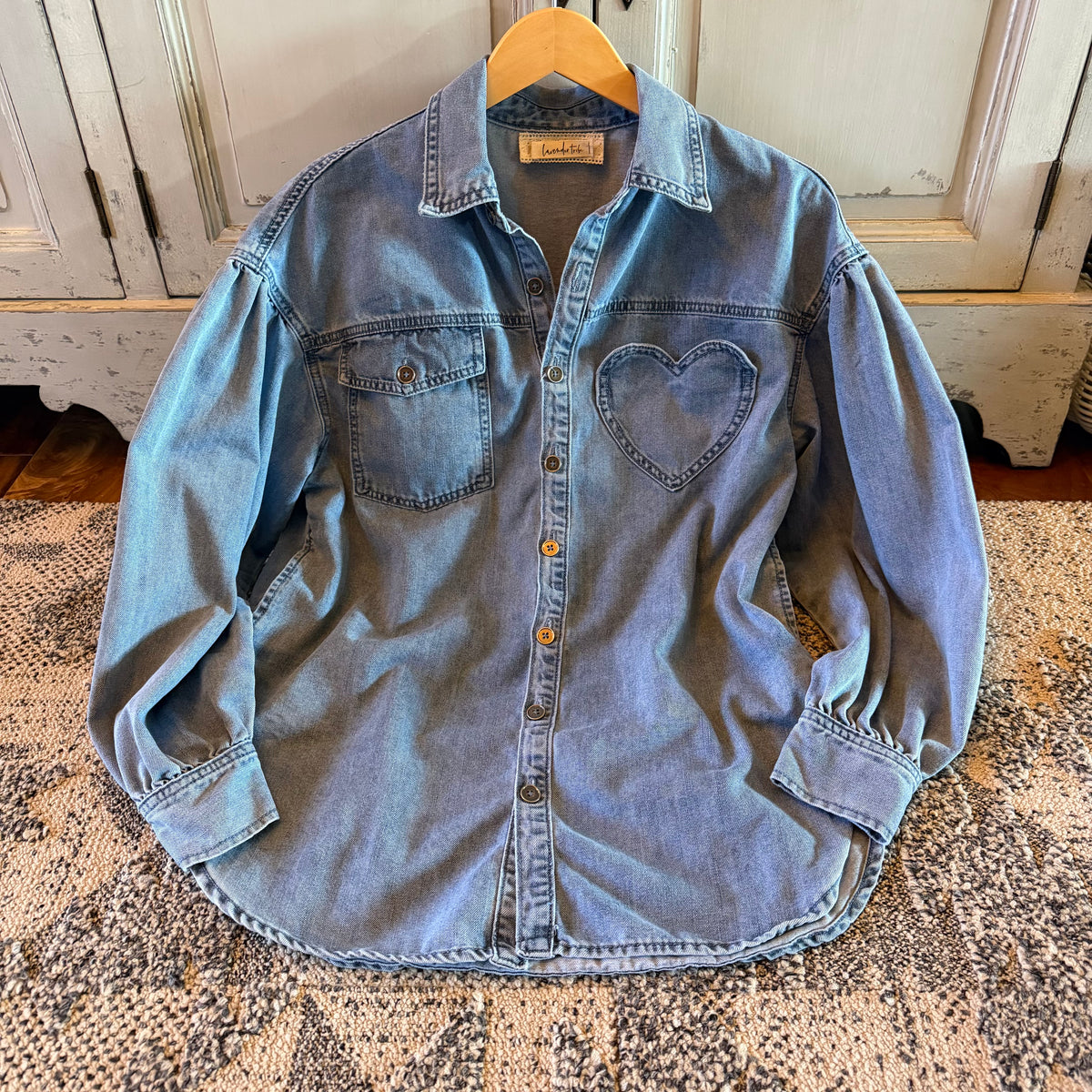 A light blue denim button-up shirt with a heart-shaped patch on the left chest, hanging on a wooden hanger on a patterned rug in front of a rustic white cabinet.