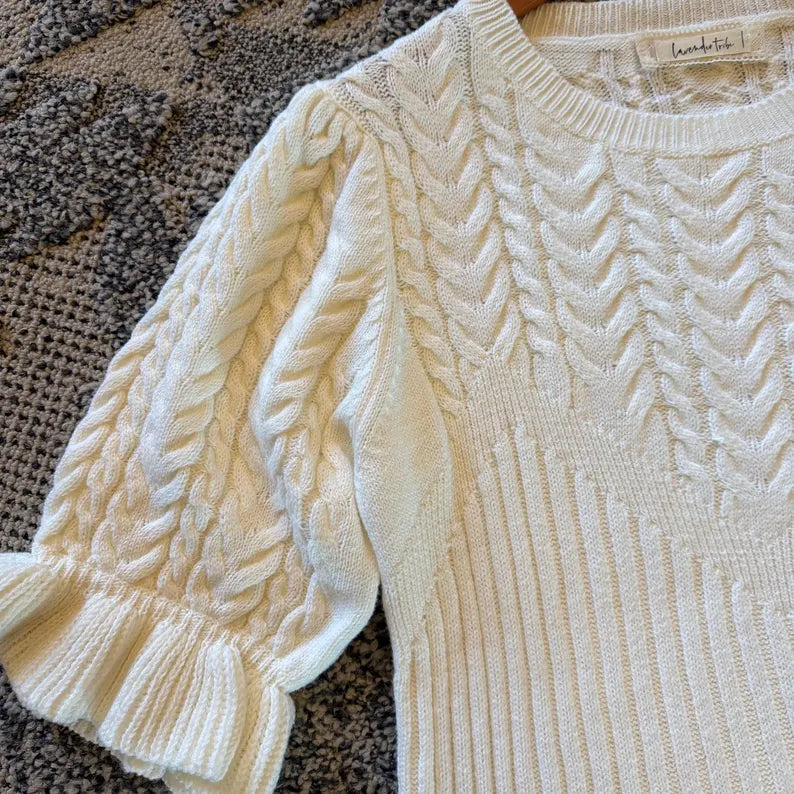 An off-white cream cable-knit short-sleeve sweater with a ruffled hem and textured chevron patterns, laid flat on a textured gray blanket. The brand name 'lambie' is visible on the inner neck label.