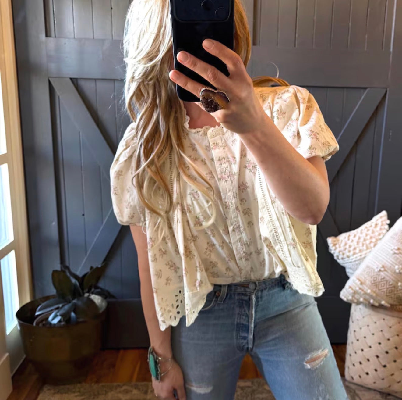Person taking a mirror selfie wearing a floral blouse and jeans in a room with a plant and decorative pillows.
