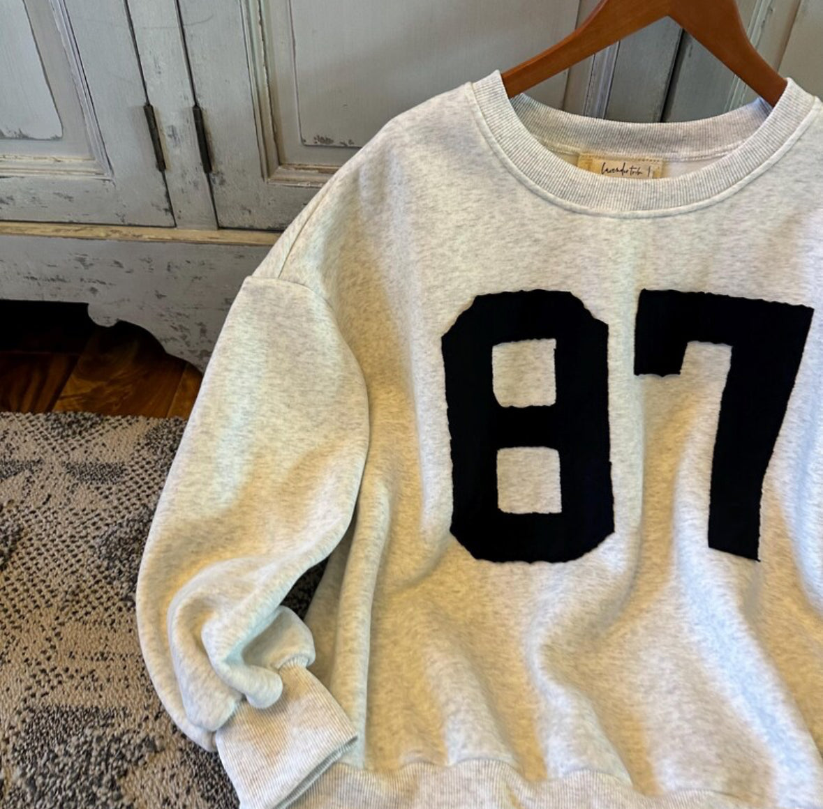 White sweatshirt with black numbers '87' on a wooden hanger against a neutral background