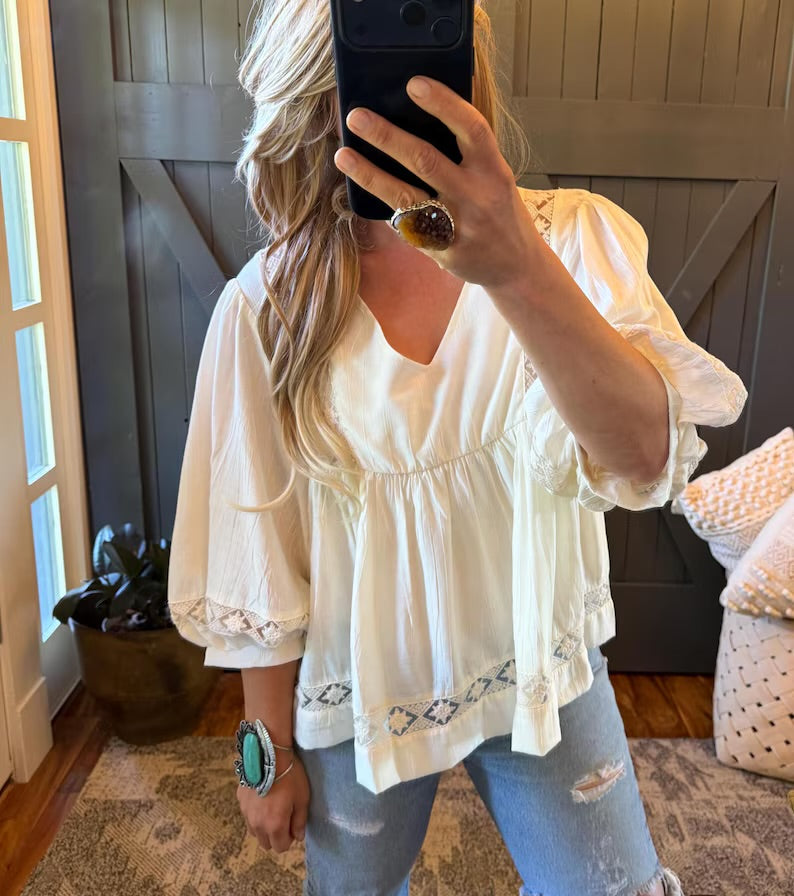 A woman takes a mirror selfie wearing a cream-colored puff-sleeve blouse with embroidered details at the hem, paired with light-wash distressed jeans.