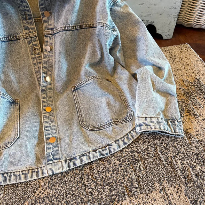 A light-wash blue denim jacket laid flat on a speckled rug, showing the front button closure with brass buttons and a chest pocket.
