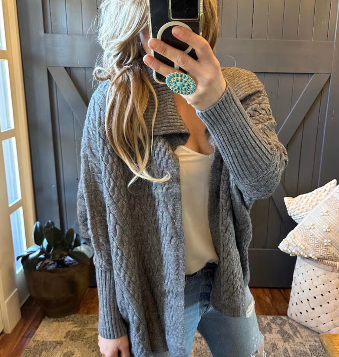 Person wearing a gray cable knit cardigan in a cozy room with wooden walls and a plant.