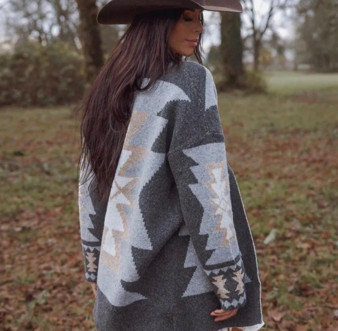 Woman wearing a patterned sweater and wide-brimmed hat in an outdoor setting with trees and grass.