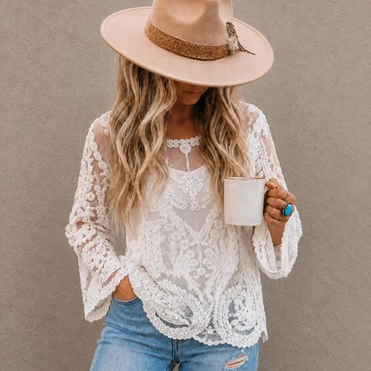 A woman wearing a beige wide-brim hat, a white lace long-sleeve top over a sheer white camisole, and light blue jeans, holding a white mug against a textured gray wall.