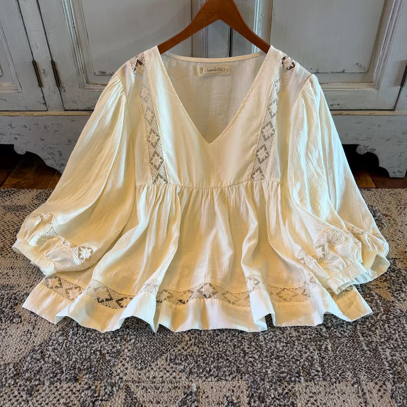 An off-white/cream bohemian-style blouse with lace trim and ruffles, hanging on a wooden hanger against a rustic white paneled cabinet background.
