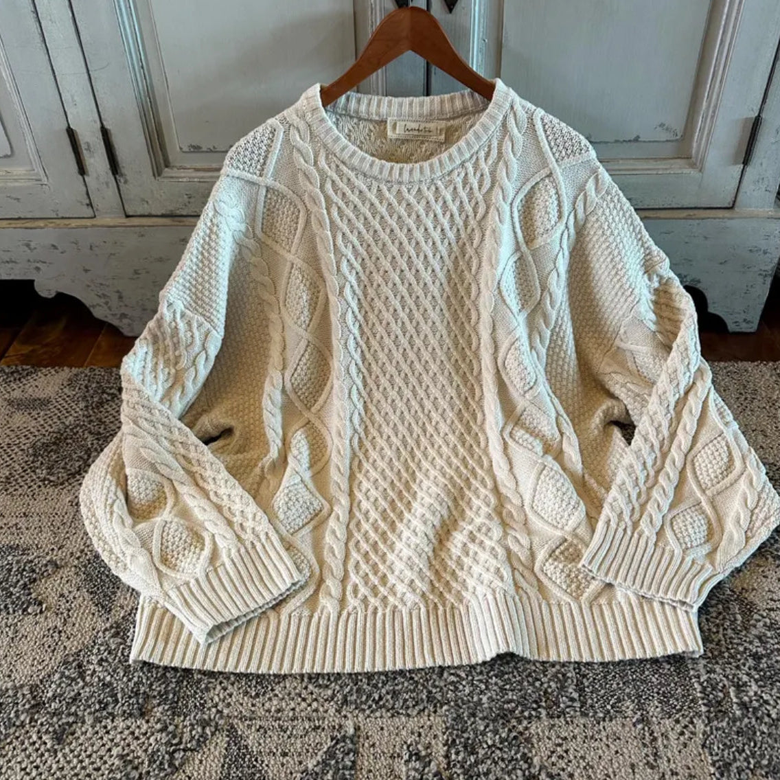 Cream-colored cable knit sweater on a wooden hanger against a rustic background