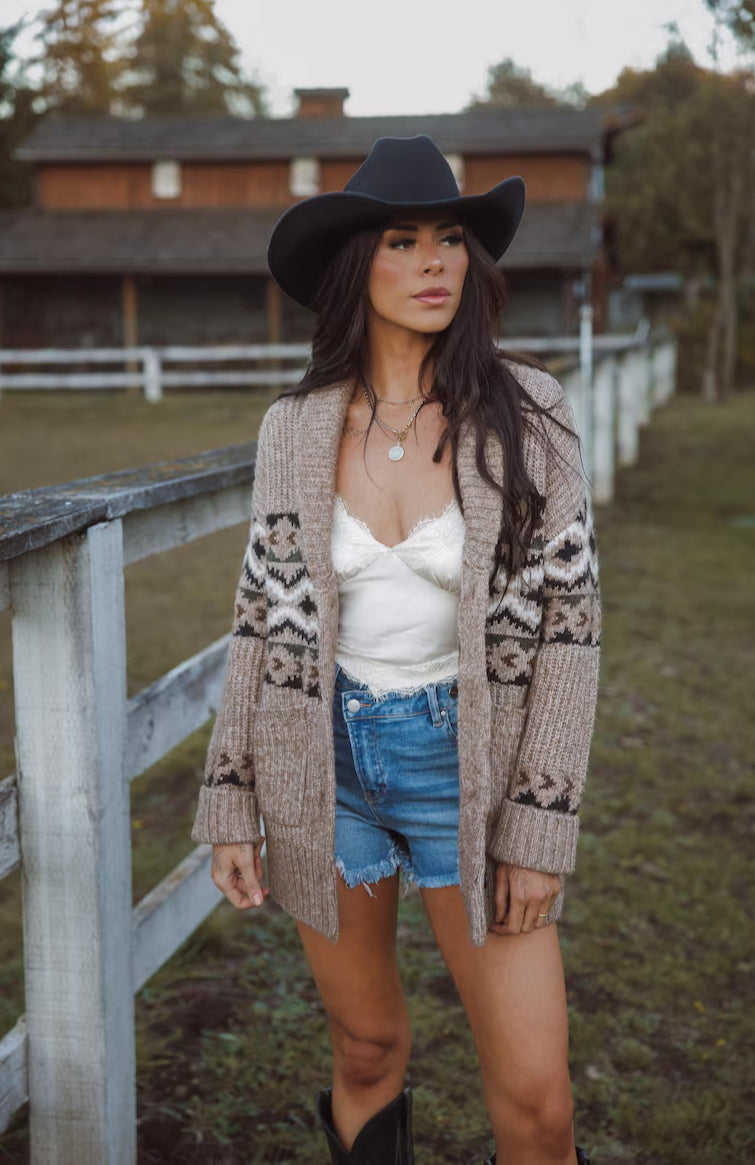 Woman wearing a patterned cardigan, denim shorts, and a cowboy hat outdoors.