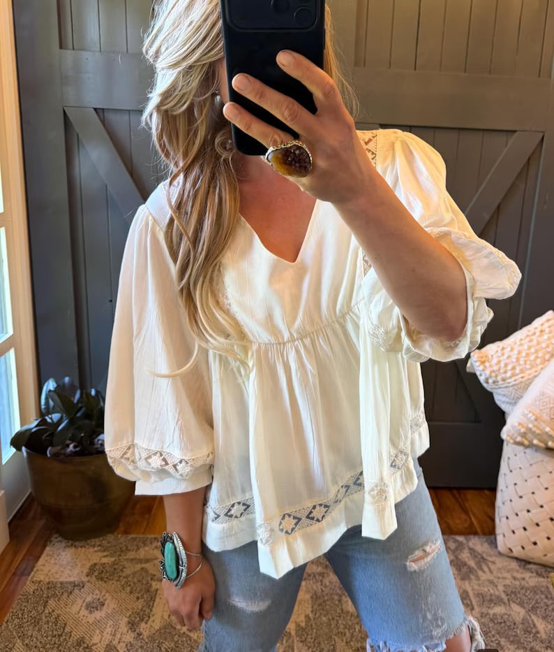 A woman takes a mirror selfie wearing a cream-colored bohemian puff-sleeve blouse with lace trim and light-wash distressed jeans.