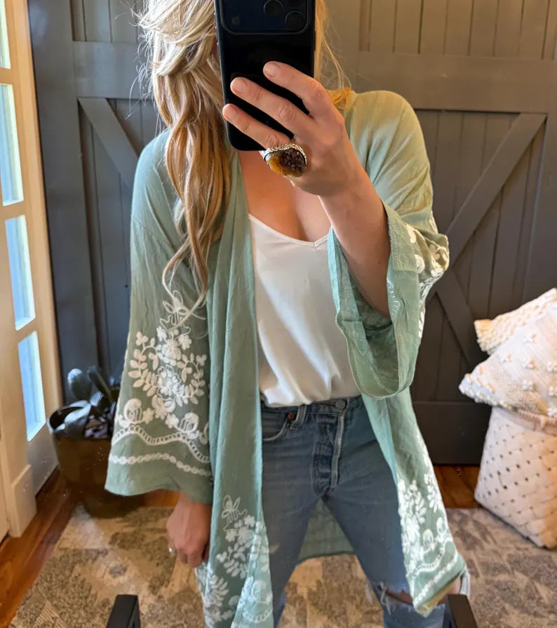 A woman takes a mirror selfie showing a sage green open-front kimono cardigan with white floral lace embroidery on the sleeves and hem, worn over a white tank top and blue jeans.