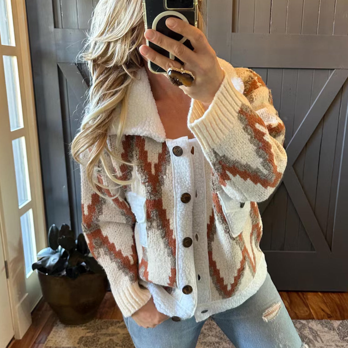 Person wearing a patterned cardigan taking a mirror selfie indoors.