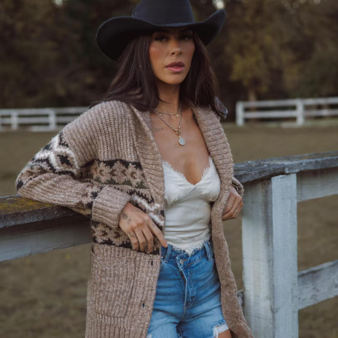 Woman wearing a patterned cardigan, white top, and blue jeans with a black cowboy hat in an outdoor setting.