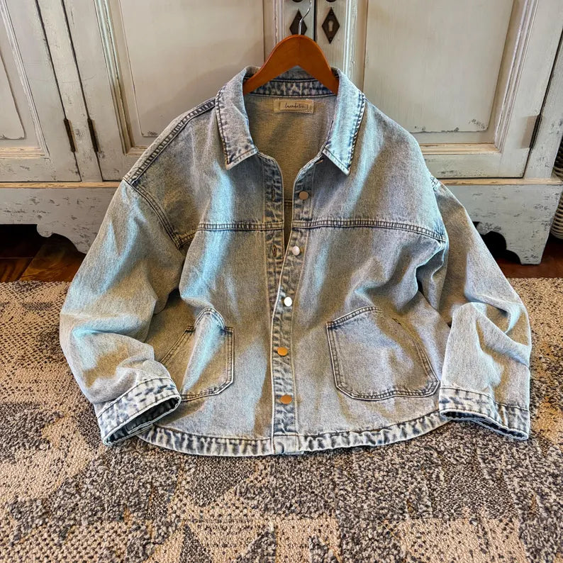 A light-wash denim jacket hanging on a wooden hanger, displayed flat on a patterned rug in front of a distressed white cabinet.