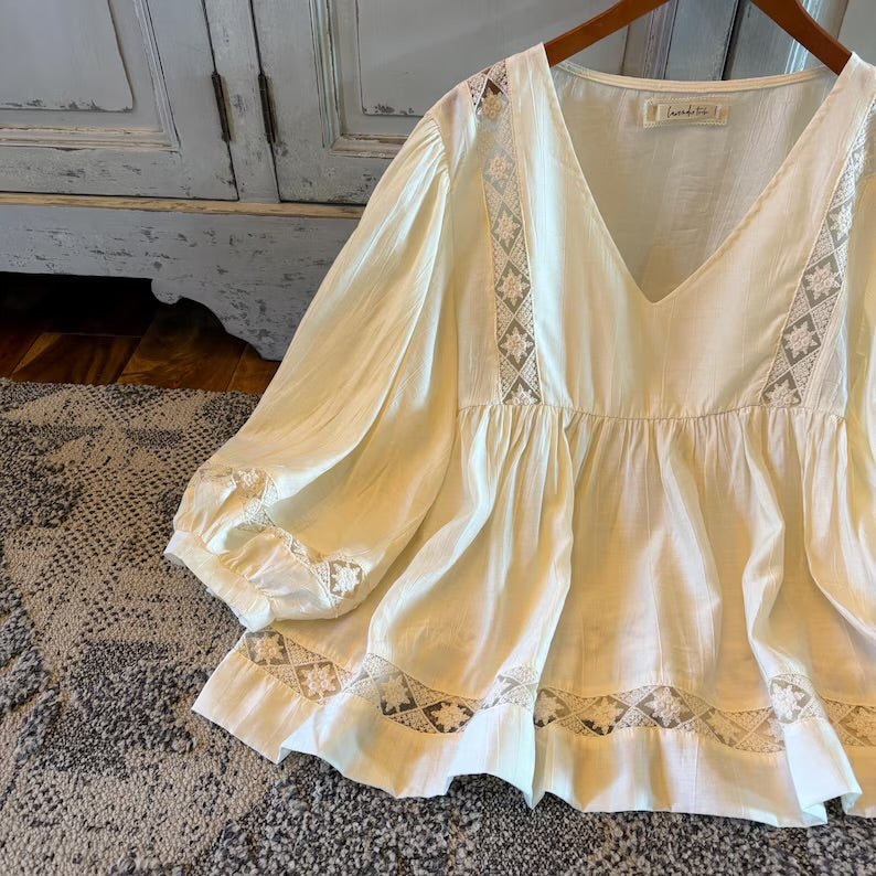 An off-white/cream blouse with long puff sleeves and geometric lace embroidery, hanging on a wooden hanger on a patterned rug in front of a distressed gray cabinet.
