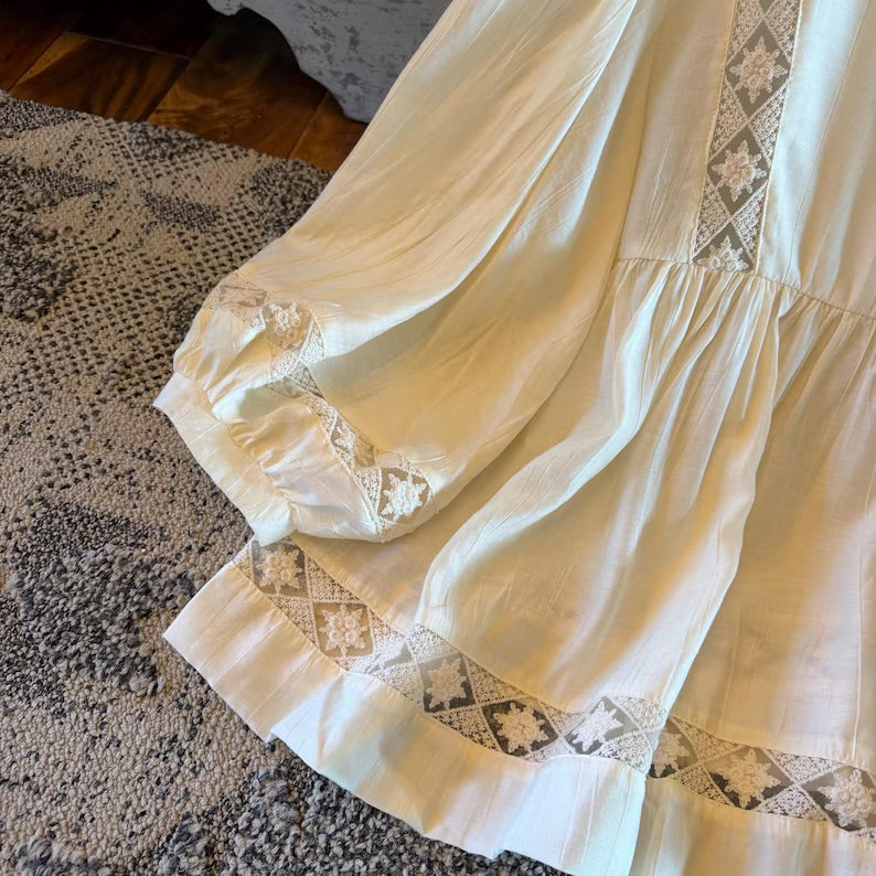A close-up of a cream-colored bohemian-style dress or long skirt with intricate geometric lace embroidery and ruffled trim, laid out on a textured gray rug.