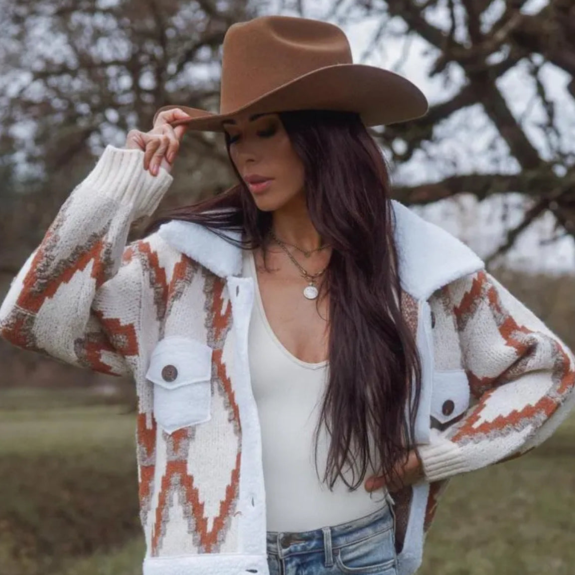Woman wearing a patterned cardigan, white top, and brown cowboy hat outdoors.