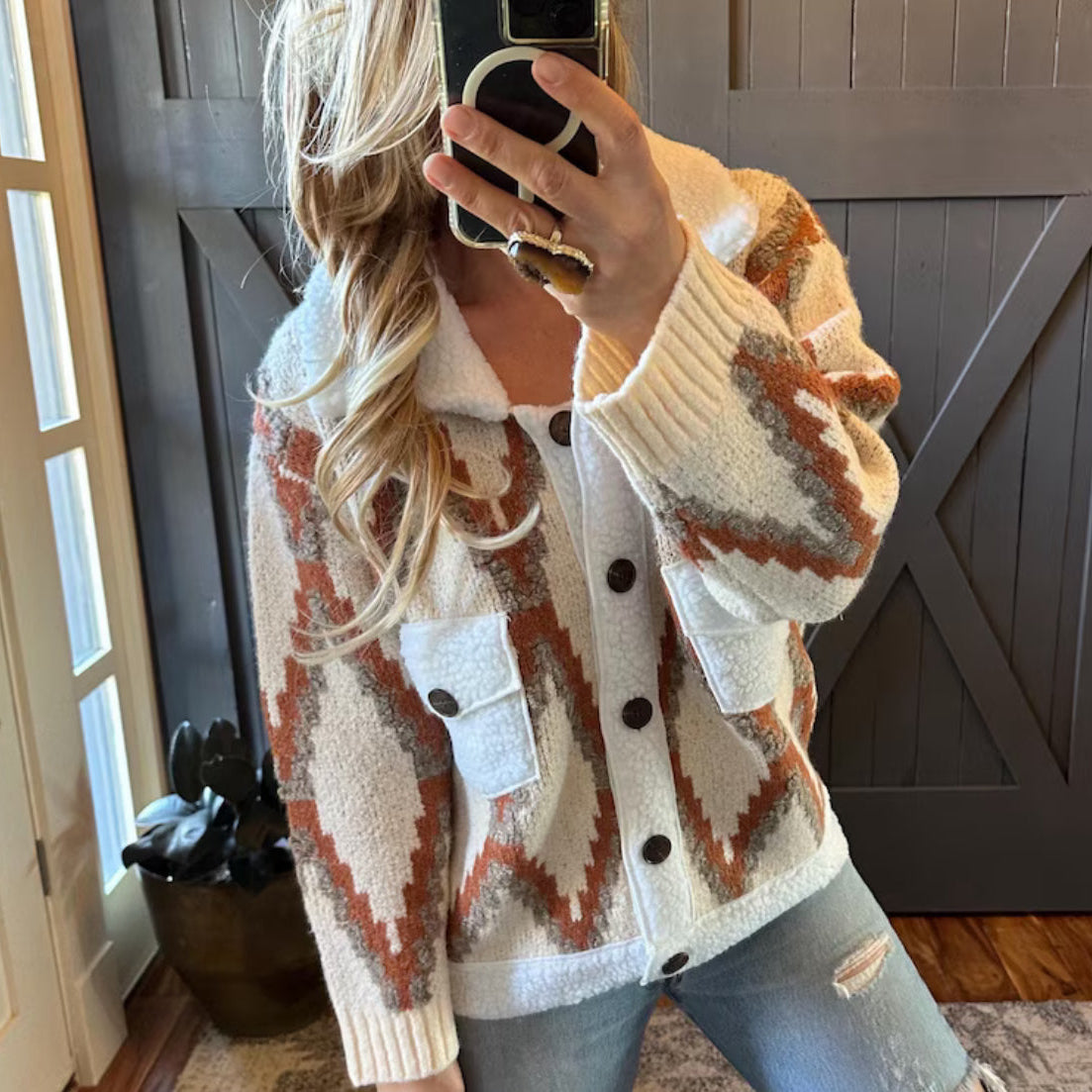 Person wearing a patterned cardigan taking a mirror selfie indoors.