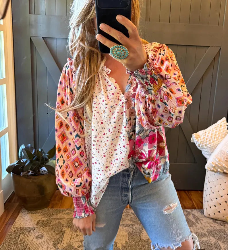 A woman takes a mirror selfie wearing a bohemian-style layered blouse with colorful geometric and floral patterns, paired with light-wash distressed jeans.