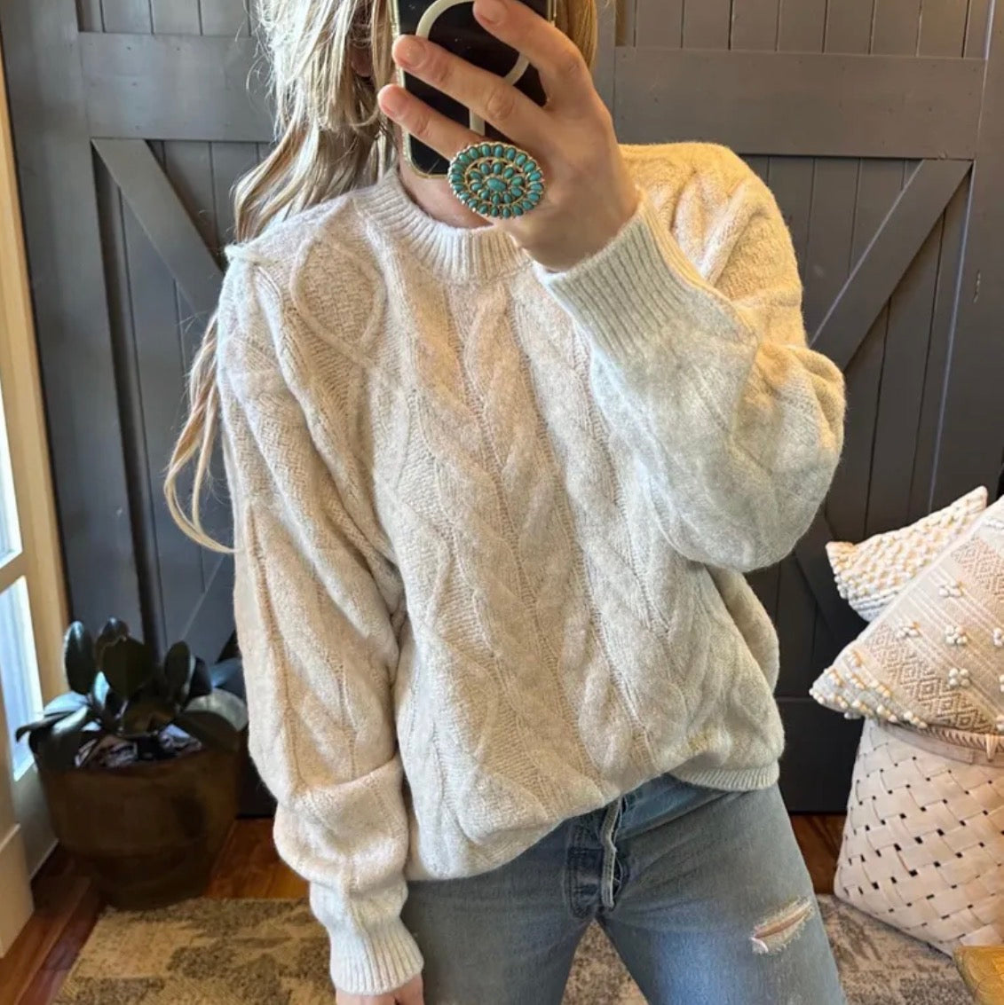 Person wearing a cream cable knit sweater and jeans, taking a mirror selfie.