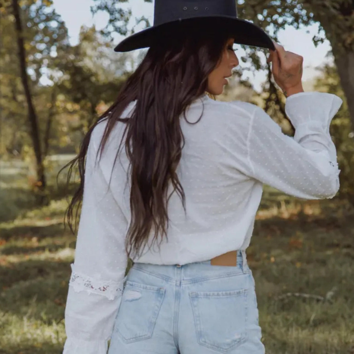 Woman wearing a white blouse and blue jeans with a black cowboy hat in a natural setting.