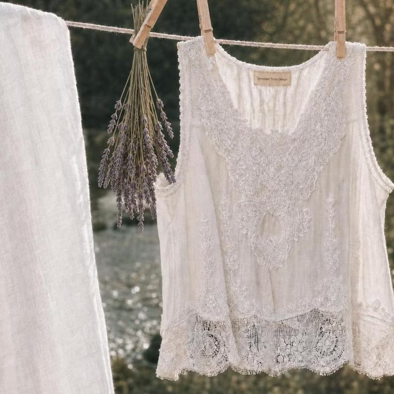 An off-white lace sleeveless blouse hanging on a clothesline with wooden clothespins, alongside a matching off-white fabric panel and a bunch of dried lavender, set against a blurred outdoor background.