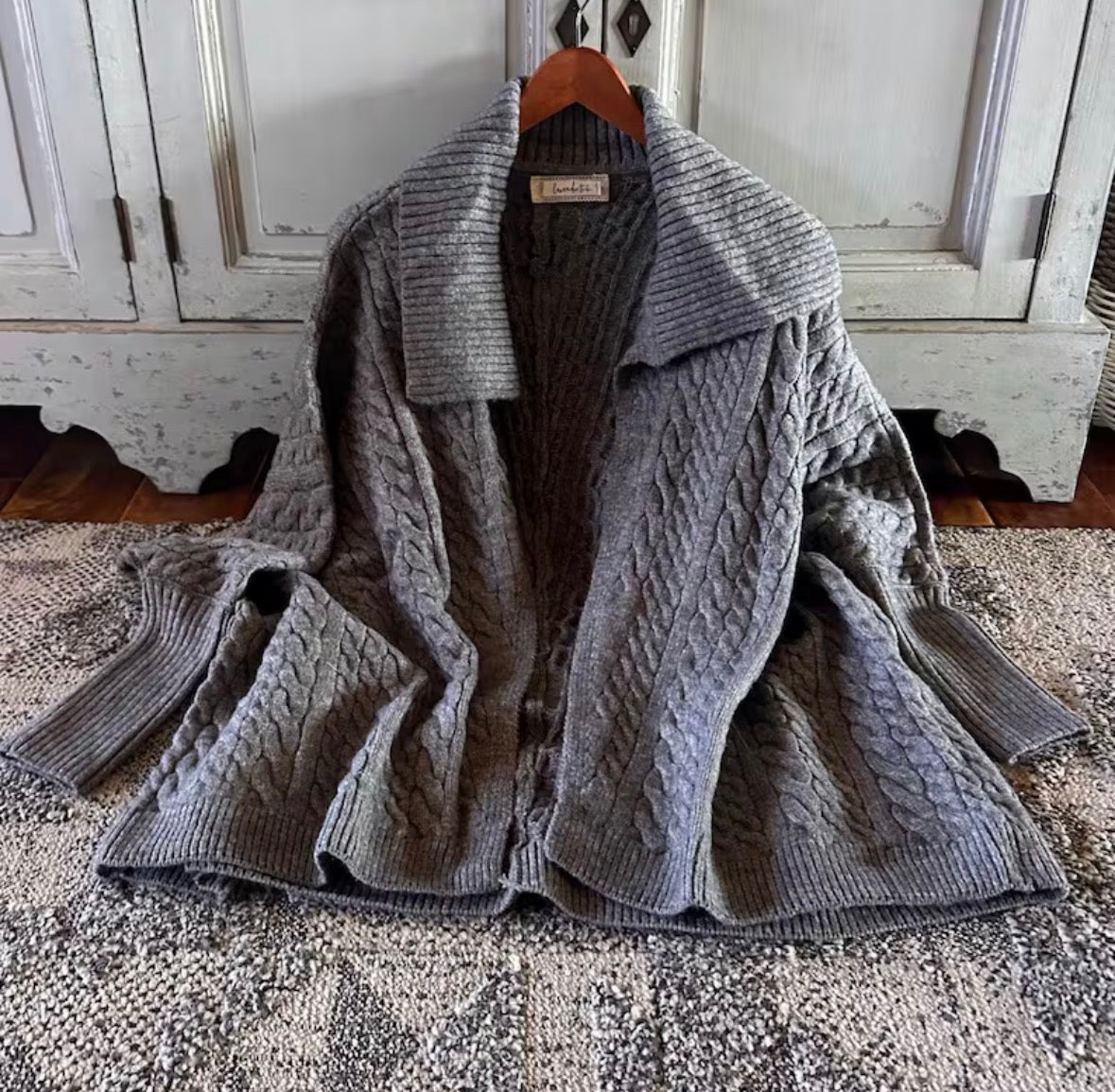 Gray cable knit cardigan on a hanger against a rustic wooden cabinet background