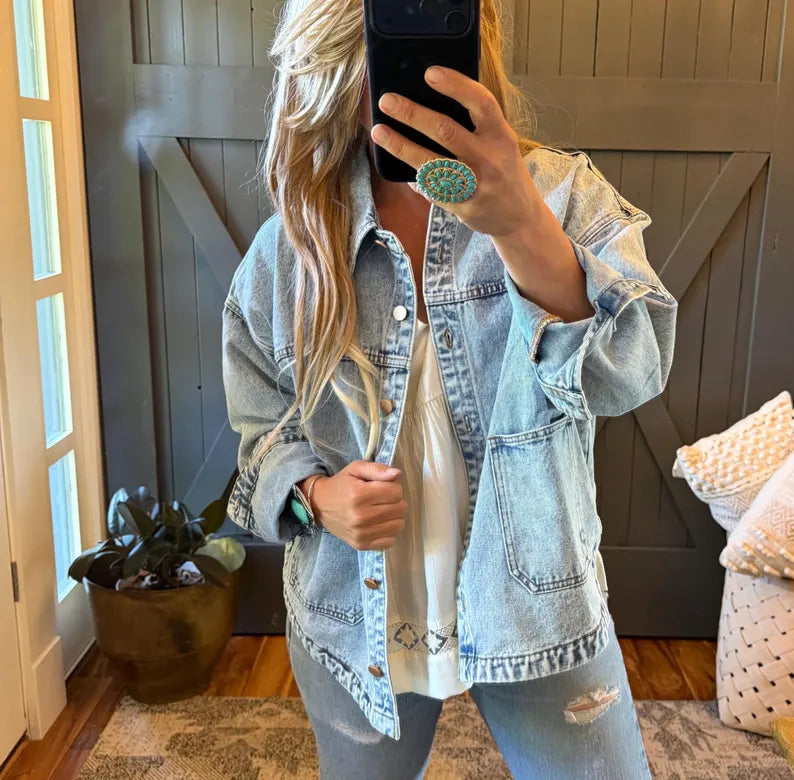 A woman takes a mirror selfie wearing a light-wash blue denim jacket over a white top and matching distressed jeans. She is standing in front of a dark gray barn-style door with a potted plant and pillows visible in the room.