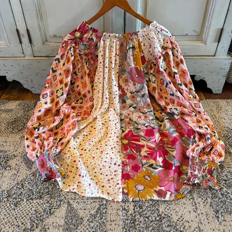 A long-sleeve bohemian-style blouse with a gathered elastic neckline, displayed on a wooden hanger over a patterned rug. The blouse features a vertical split design with one side in a pink and orange geometric print and the other in a vibrant floral print with pink, yellow, and red flowers.