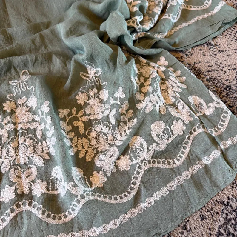 A sage green fabric textile with intricate white floral embroidery and lace trim, draped on a speckled stone surface.