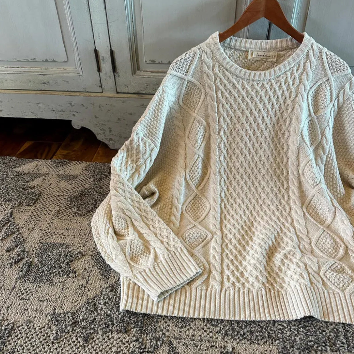 Beige cable knit sweater on a wooden hanger against a rustic wooden cabinet background.