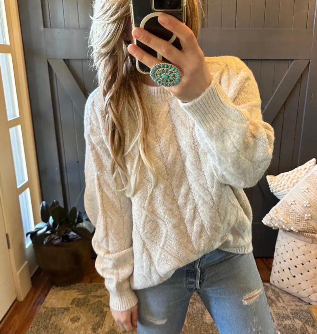 Person wearing a cream cable knit sweater and jeans, taking a mirror selfie.