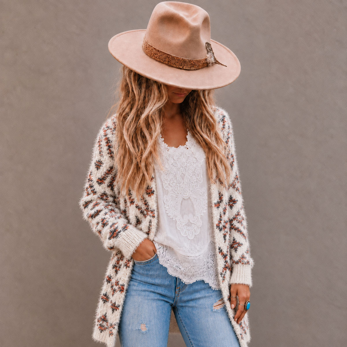 A woman wearing a cream knit cardigan with brown and rust floral patterns, a white lace top, blue jeans, and a beige wide-brim hat, posing against a neutral gray wall.