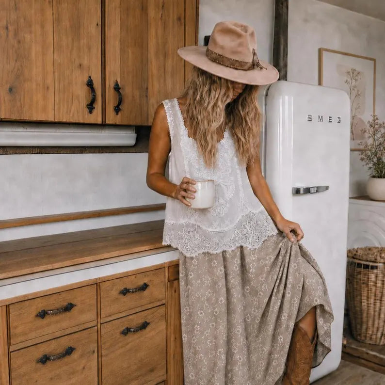 A woman stands in a rustic kitchen wearing a beige wide-brim hat, a white sleeveless lace top, and a beige patterned maxi skirt, holding a white mug next to a white retro SMEG refrigerator.