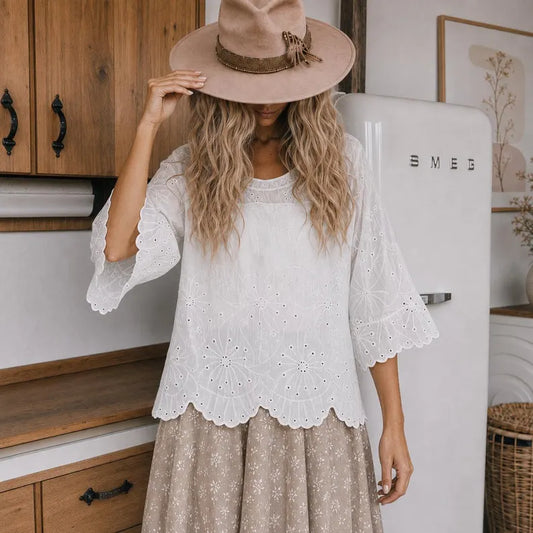 A woman wearing a bohemian-style outfit in a kitchen setting, featuring a white eyelet lace top with bell sleeves, a beige hat, and a beige patterned skirt.