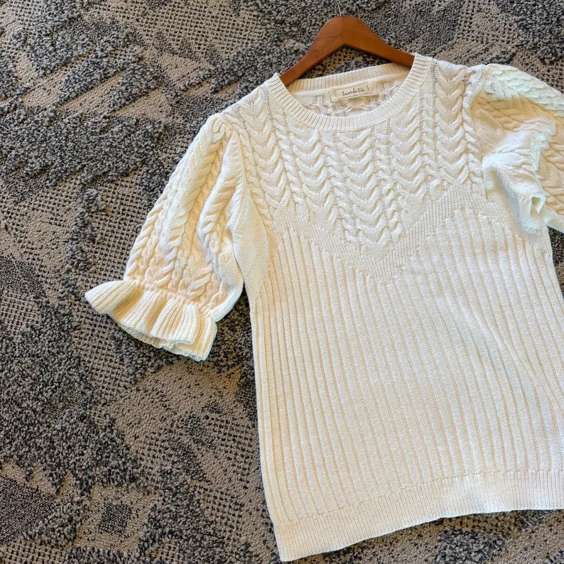 An off-white/cream cable-knit short-sleeve sweater with textured sleeves and a ribbed hem, displayed on a wooden hanger over a gray patterned rug.